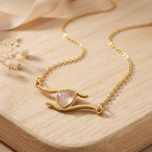 Gros plan du collier Milos, cœur en quartz rose entre deux vagues dorées, sur un support en bois - LELIEGE 